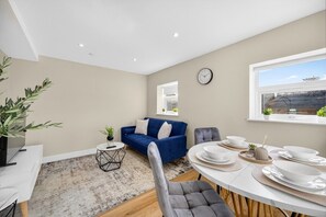 A modern living room with a blue sofa, a dining table set with white plates, a small side table with a plant, and a large window with a view. - 2BR - Fast Wi-Fi - Work Space - Free Parking (Cardiff)