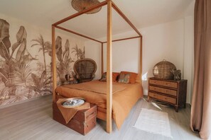 A room with a four-poster bed, a large mural, a wicker chair, a wooden dresser, and a suitcase.