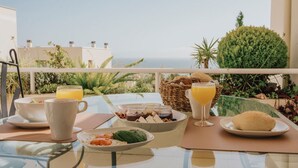 Un plateau de petit-déjeuner installé sur un balcon avec vue sur la mer, comprenant une table avec des assiettes, des tasses et un panier de pain.