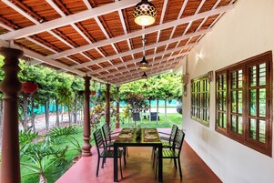 Área de refeições coberta ao ar livre, com mesa e cadeiras, rodeada por vegetação e vista para um jardim paisagístico.