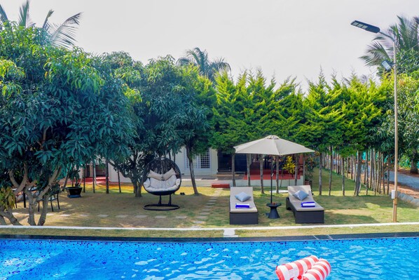 A swimming pool with lounge chairs and a hanging chair, surrounded by lush greenery and a covered seating area. - Alaya Stays Eco Serenity 2-BHK With Pool (Doddaballapur)