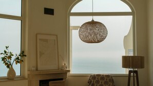 Une pièce dotée d’une grande fenêtre en arc de cercle, d’un luminaire suspendu en fibres tressées, d’une petite table recouverte d’une nappe à motifs et d’un vase avec des plantes posé sur le rebord de la fenêtre.