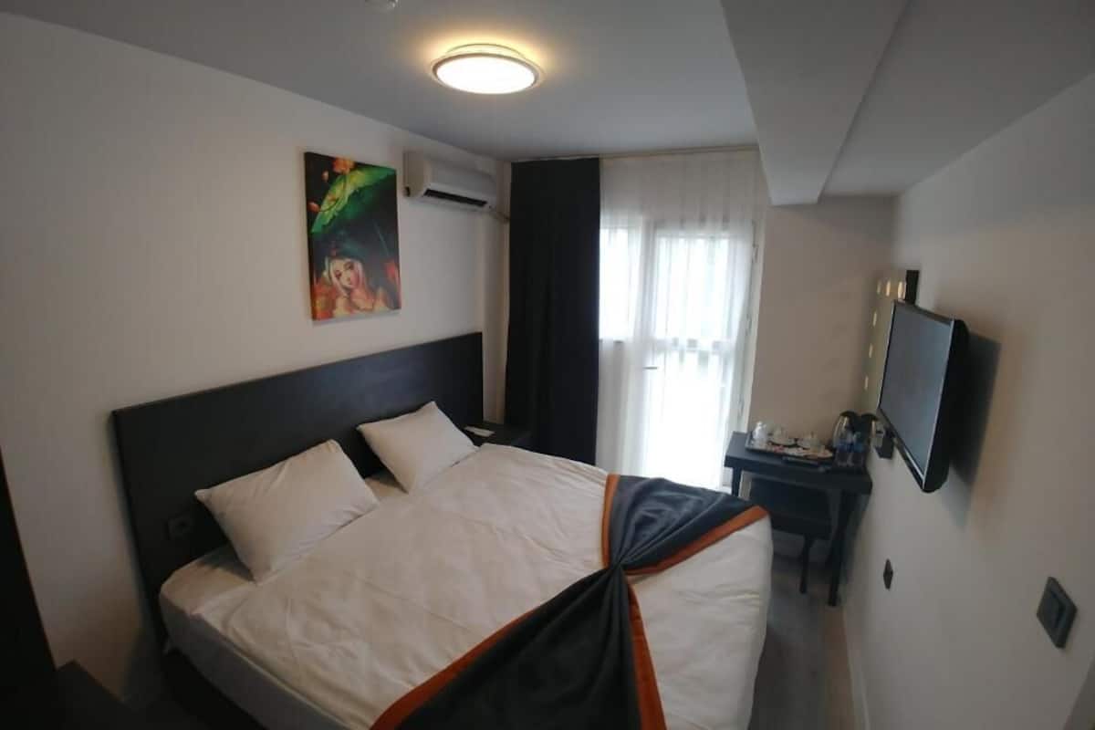 Habitación de hotel con cama, un cuadro en la pared, escritorio con televisor y ventana con cortinas.