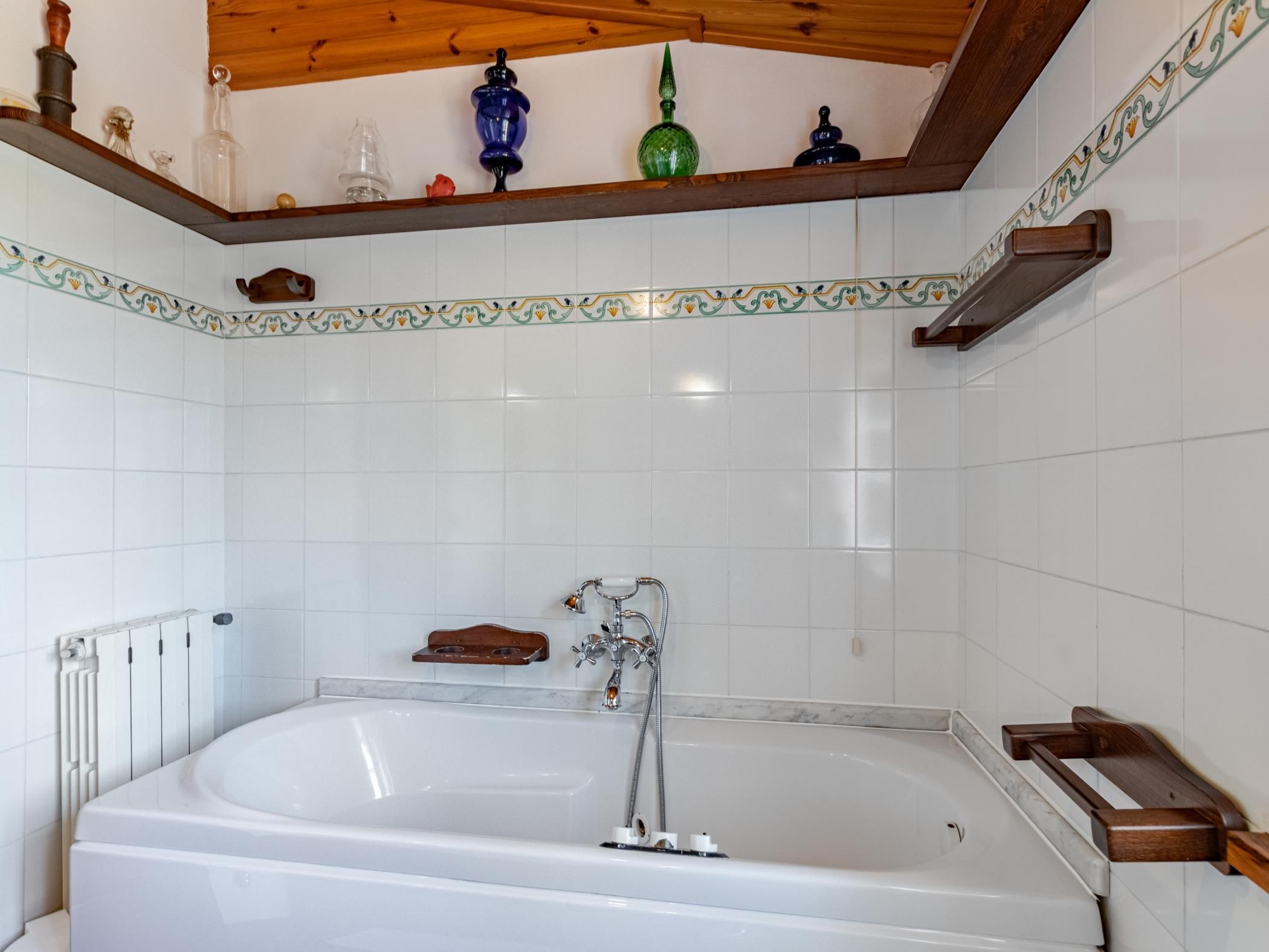 A bathroom with a white bathtub, a wooden shelf with decorative items, and a tiled wall with a decorative border.