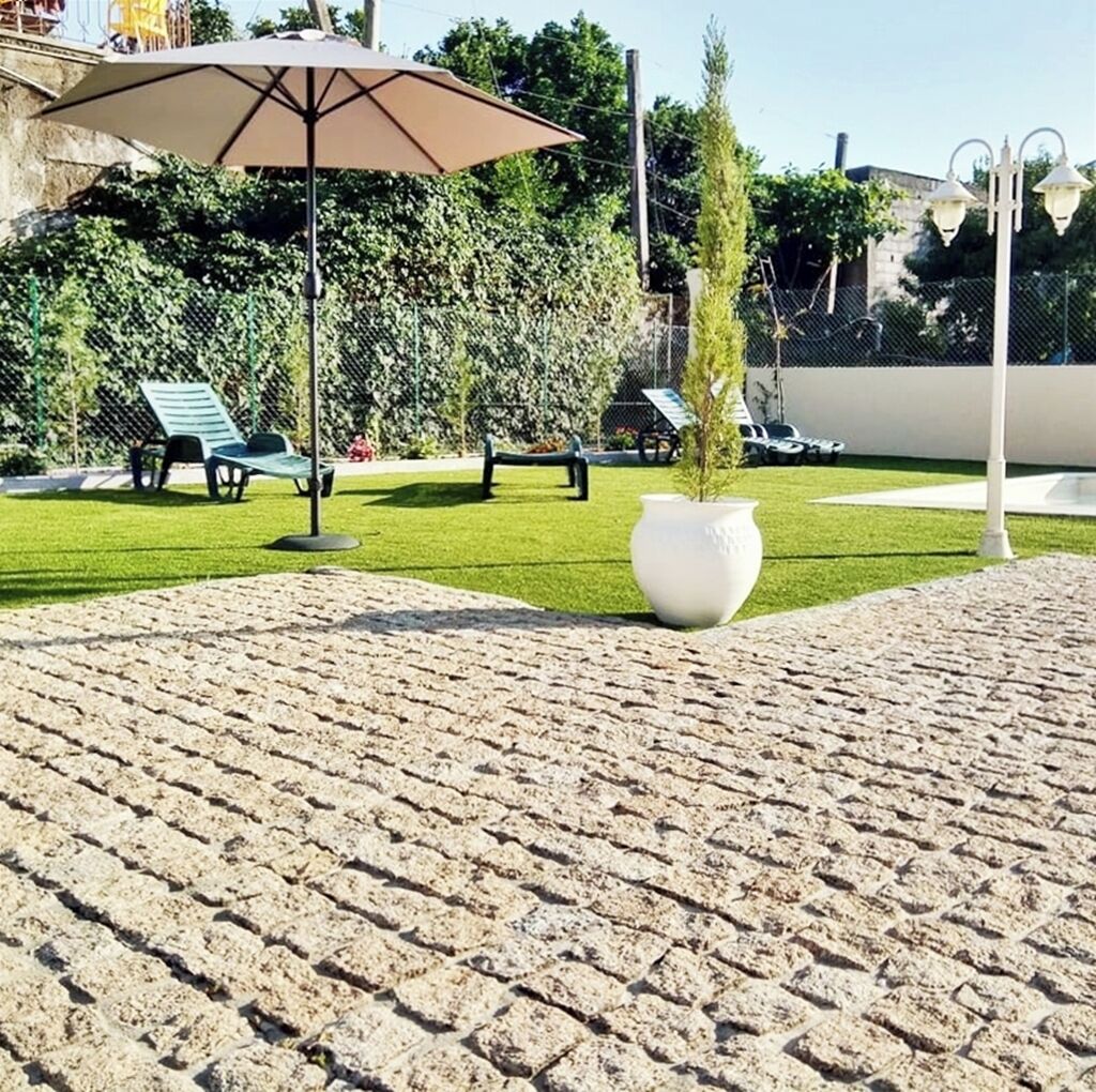 A paved outdoor area with a large white planter, green lounge chairs, and a brown umbrella.