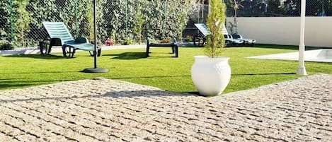A paved outdoor area with a large white planter, green lounge chairs, and a brown umbrella.