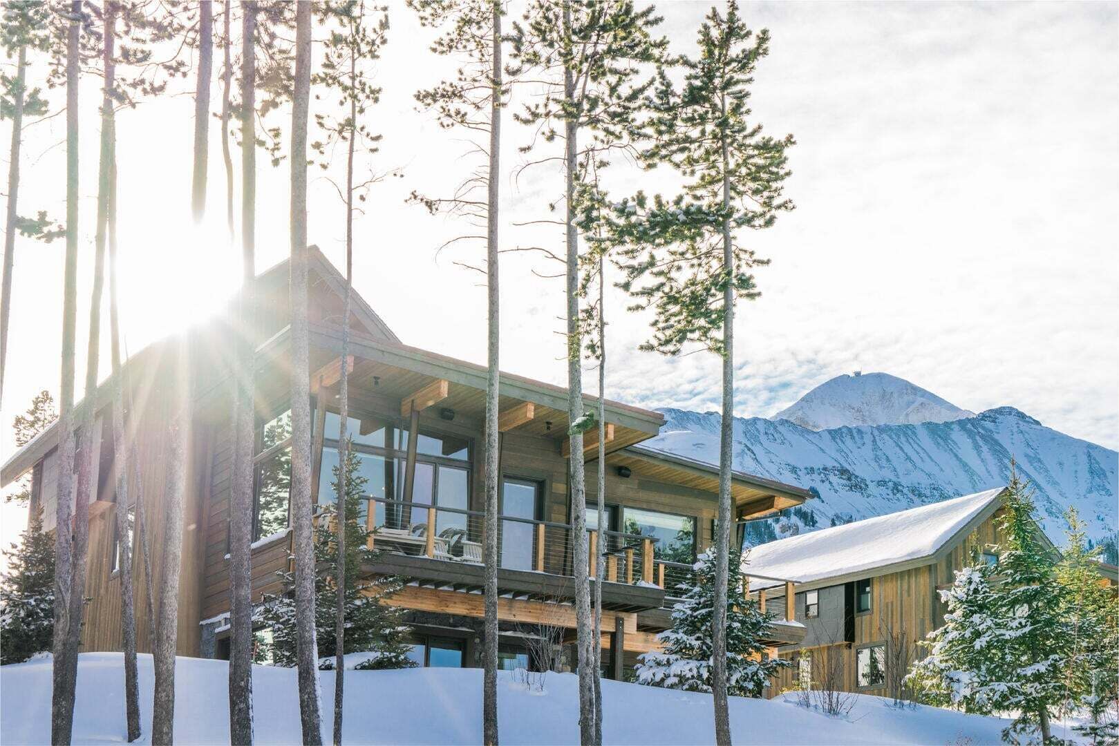 Un chalet moderne en bois, doté de grandes fenêtres et d’un balcon, entouré de grands arbres et surplombant un paysage montagneux enneigé.