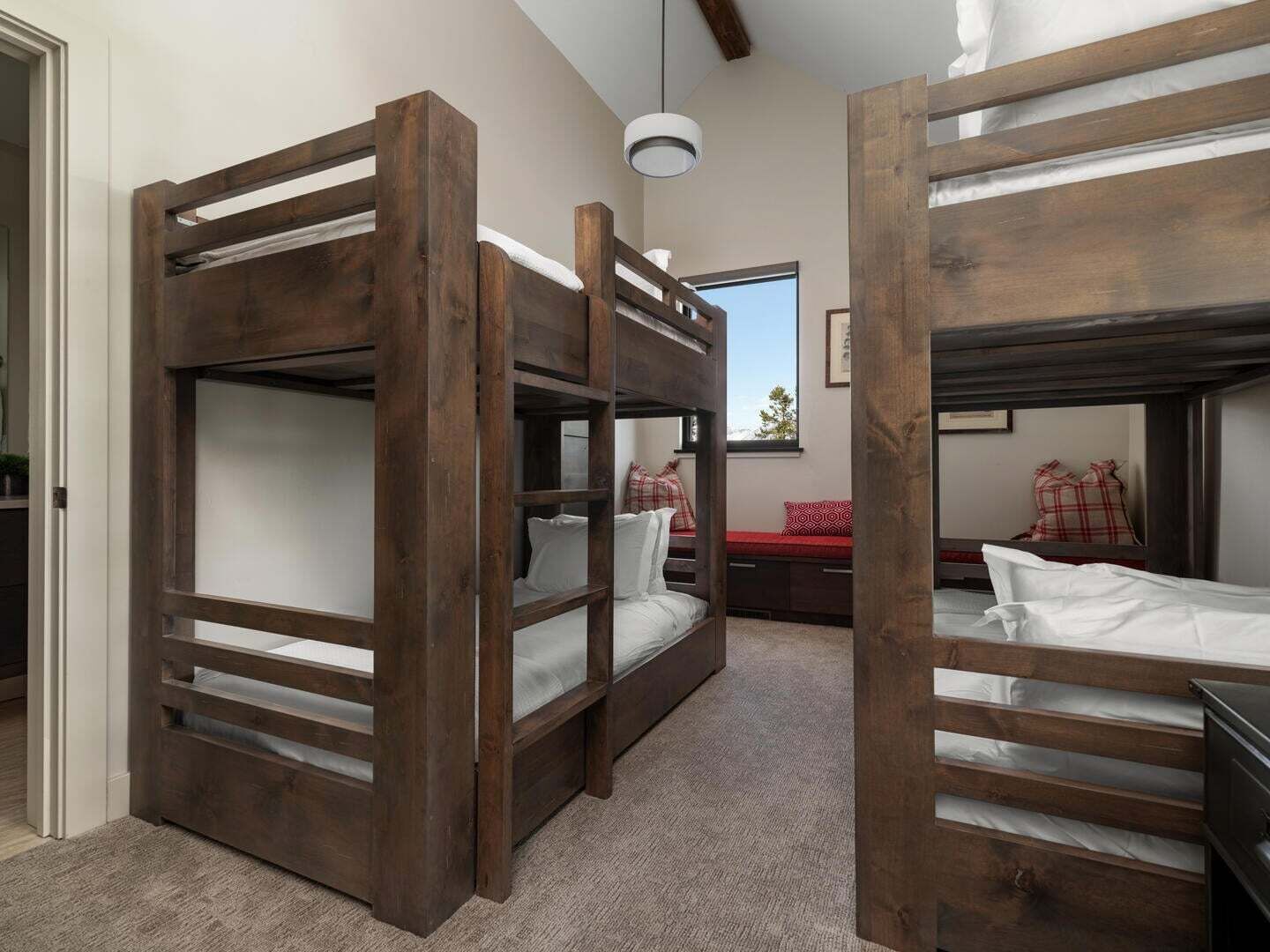 Une pièce avec trois lits superposés en bois foncé, un petit bureau agrémenté d’un coussin à motifs rouges et blancs, et une fenêtre donnant sur le ciel.