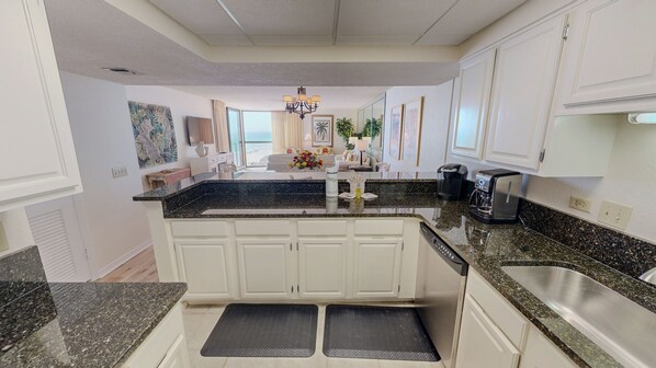 A kitchen with white cabinets, granite countertops, a built-in dishwasher, and a coffee maker. - Mainsail 152 2 Bedroom Condo (Miramar Beach)