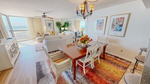 A spacious living area with a large window overlooking the beach, a dining table with chairs, a ceiling fan, and a chandelier. - Mainsail 152 2 Bedroom Condo (Miramar Beach)
