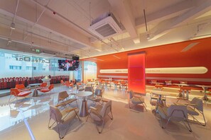 A modern dining area with tables and chairs, a large screen displaying an event, and a bright orange accent wall. - SilMiCore Hostel (Shanghai)