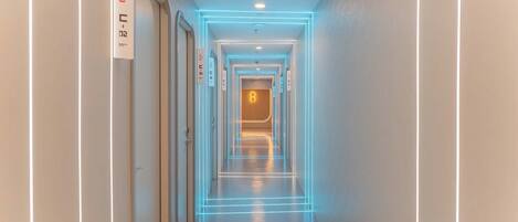 A modern hotel corridor with illuminated lines on the walls, a visible elevator, and an emergency exit sign.