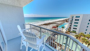 A balcony with white chairs, a view of a beach, a pool area, and a building with multiple windows. - Islander Beach Resort 705 Condo (Fort Walton Beach)