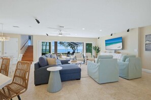 A spacious living room with a large sofa, a round coffee table, and a TV mounted on the wall. There is a staircase leading to another level, and large windows offering a view of the outdoors. - Nst108 4 Bedroom Home (Punta Gorda)