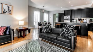 Un salon moderne avec un canapé en cuir noir, des coussins à motifs, une table basse en verre et un tapis à motifs géométriques.