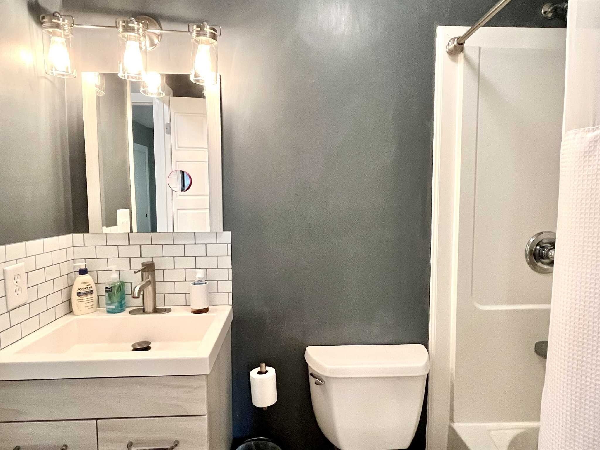 A bathroom with a white sink, mirror, and toilet. There are toiletries on the sink and a towel hanging on the door.