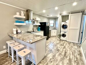 Cocina compacta con encimera de granito, taburetes blancos de bar, horno empotrado y lavadora-secadora.