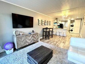 Ein modernes Wohnzimmer mit einem großen Flachbildfernseher, einem Schrank mit Fischgrätmuster und einem Couchtisch. Es gibt eine Küchenzeile mit Spüle, Herd und einer integrierten Waschmaschine. An der Wand ist das Wort „BEACH“ angebracht.