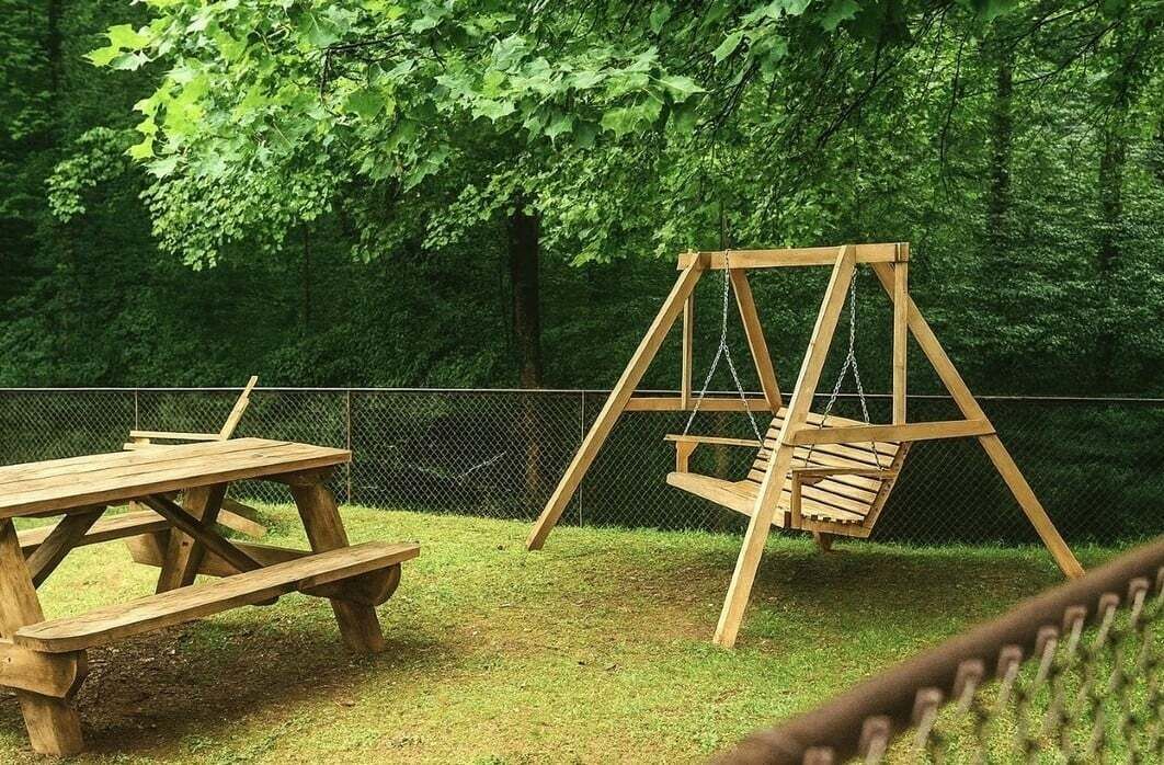 Un conjunto de columpios de madera y una mesa de picnic en un área exterior cercada, con abundante follaje verde al fondo.