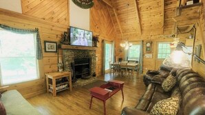 L’intérieur chaleureux d’un chalet en bois comprend une cheminée, un canapé en cuir, une table à manger en bois et un téléviseur fixé au mur au-dessus de la table.