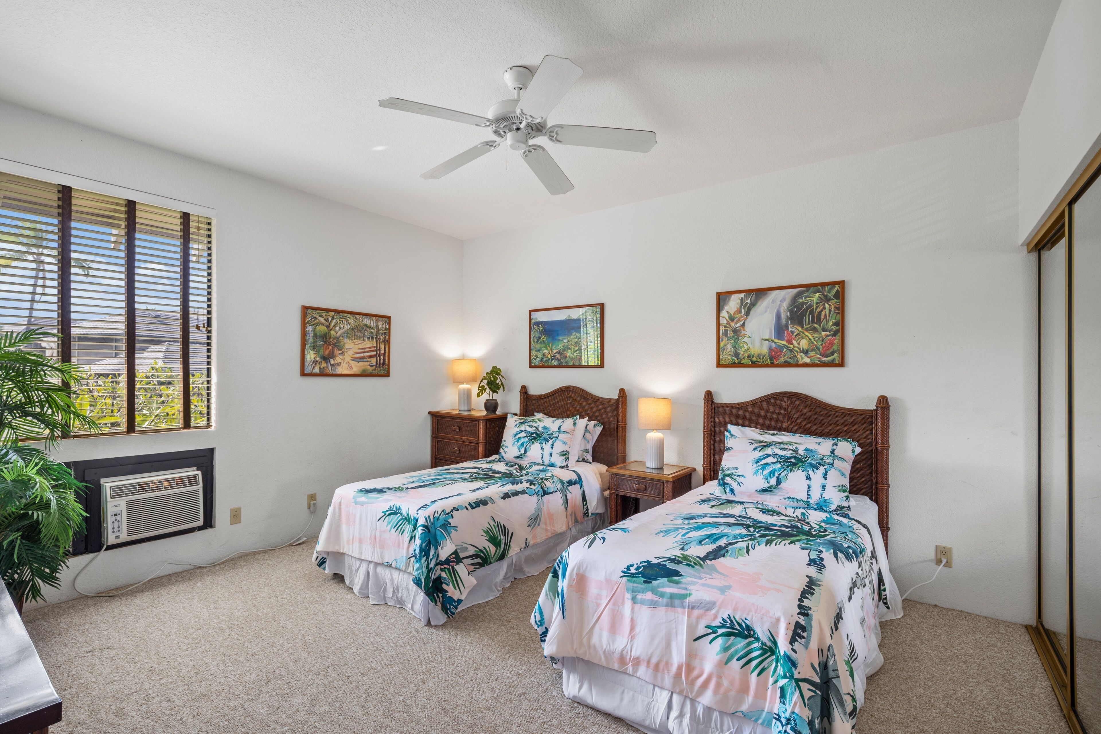 A hotel room with two beds, a ceiling fan, a window with blinds, and artwork on the walls.