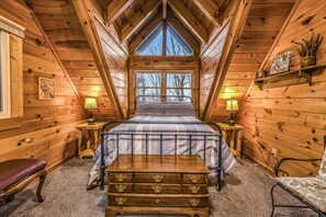 A cozy wooden cabin bedroom with a bed, bedside lamps, a dresser, and a window with a view of trees. - Rise And Shine 3 Bedroom Cabin (Gatlinburg)