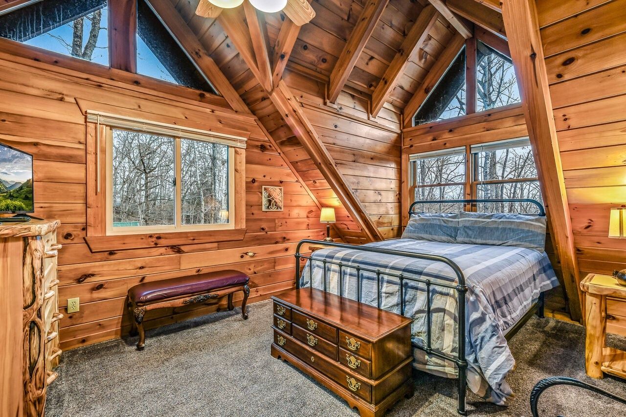 A wooden cabin bedroom with a bed, bench, dresser, and large windows.