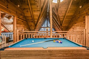A pool table in a wooden cabin with a sloped ceiling and a view of the outdoors. - Rise And Shine 3 Bedroom Cabin (Gatlinburg)