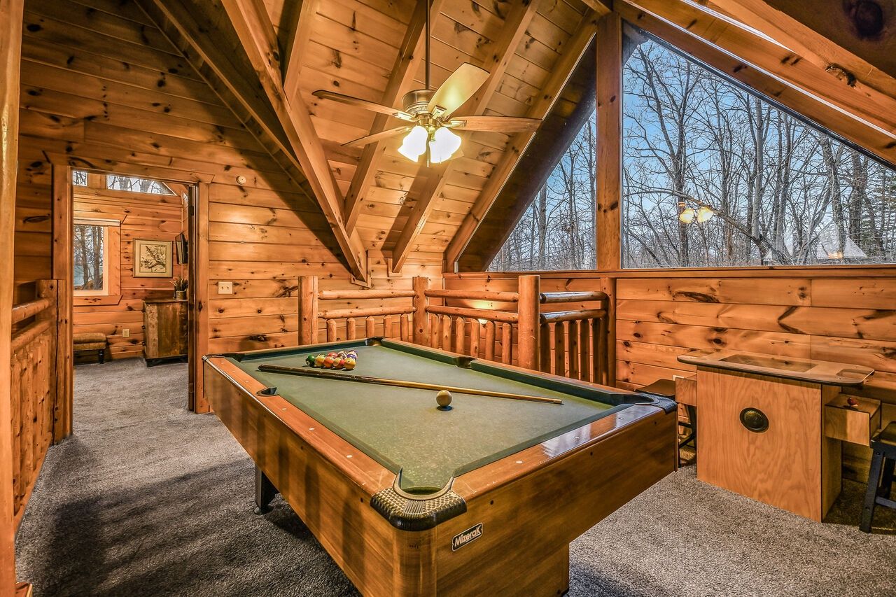 A wooden cabin interior with a pool table, a ceiling fan, and a large window offering a view of trees.