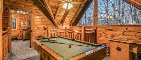 A wooden cabin interior with a pool table, a ceiling fan, and a large window offering a view of trees.