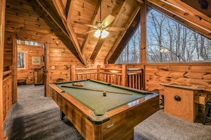 A wooden cabin interior with a pool table, a ceiling fan, and a large window offering a view of trees. - Rise And Shine 3 Bedroom Cabin (Gatlinburg)