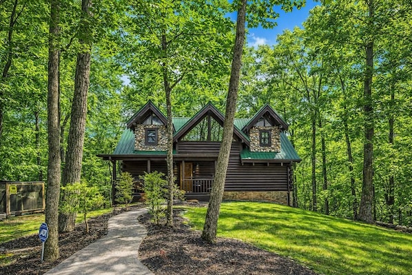 A log cabin with a green roof, surrounded by trees, and a pathway leading to the entrance. - Rise And Shine 3 Bedroom Cabin (Gatlinburg)