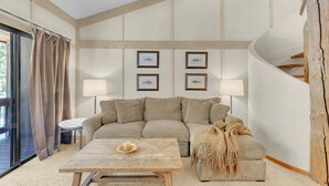 Un salon chaleureux avec un canapé, une table basse et une lampe. Les murs sont ornés de tableaux encadrés et une fenêtre est agrémentée de rideaux.