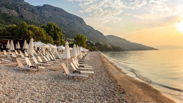 Ein Kiesstrand mit Liegestühlen und Sonnenschirmen, ruhiges Meer und Berge im Hintergrund.