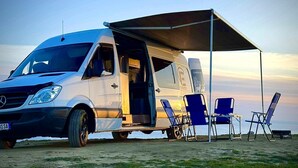 A white camper van with an open door, a canopy, and blue folding chairs on a grassy area. - Campervan Tirana Leoventure (Tirana)