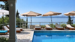 A poolside area with lounge chairs and umbrellas, a stone-tiled deck, and a view of the sea. - Hotel Barbati - Poolside Gem with Sea views (Mparmpati)