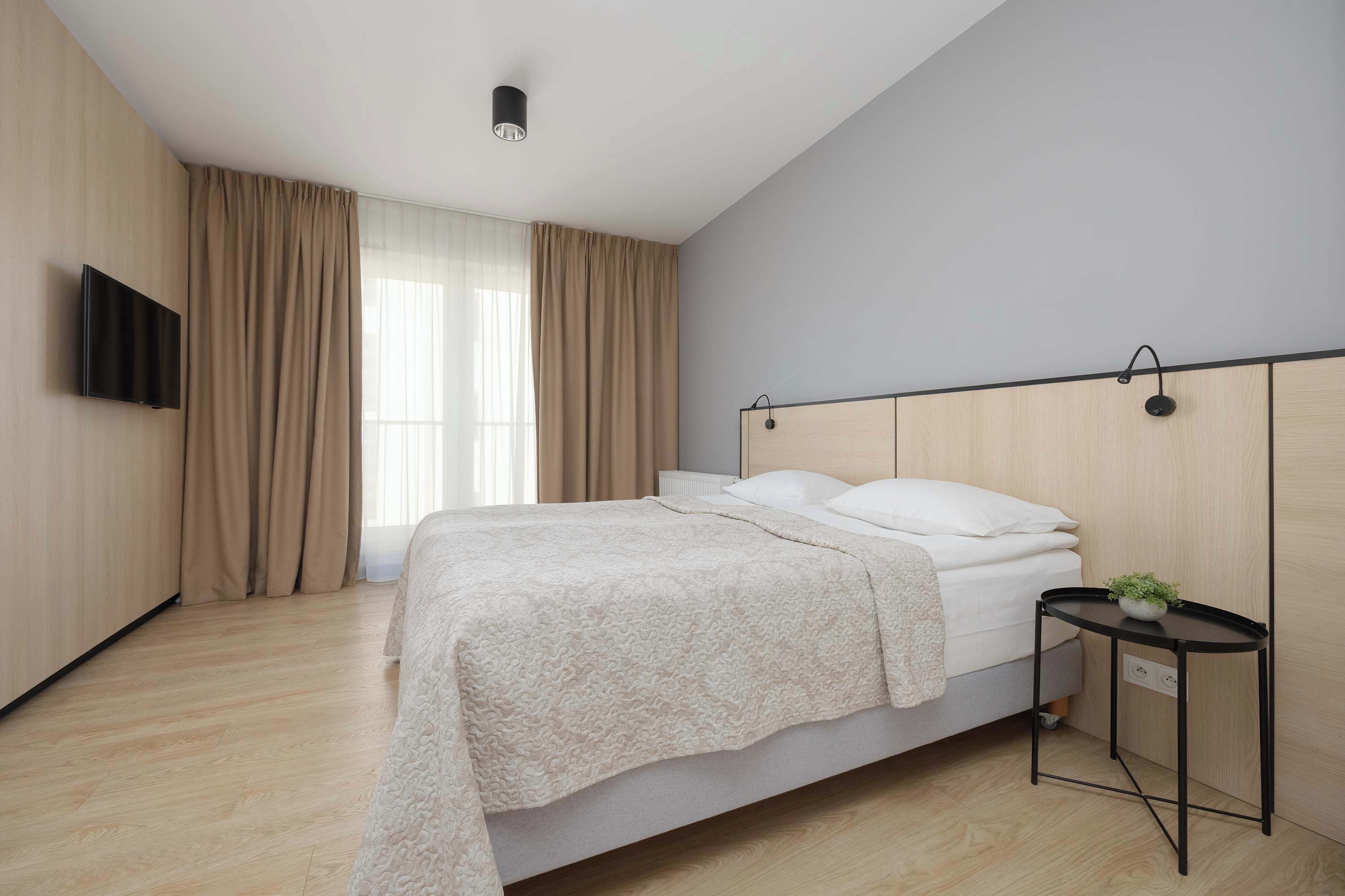 Un dormitorio moderno con cama, televisor montado en la pared, mesita de noche con planta y ventana con cortinas.