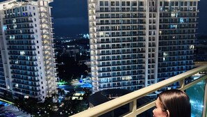 Een vrouw met een glas op een balkon, met uitzicht op hoge gebouwen en een zwembad.