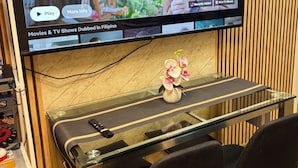 Een televisie waarop een programma met ondertiteling in een vreemde taal wordt vertoond, een glazen tafel met stoelen, en een decoratieve orchidee.