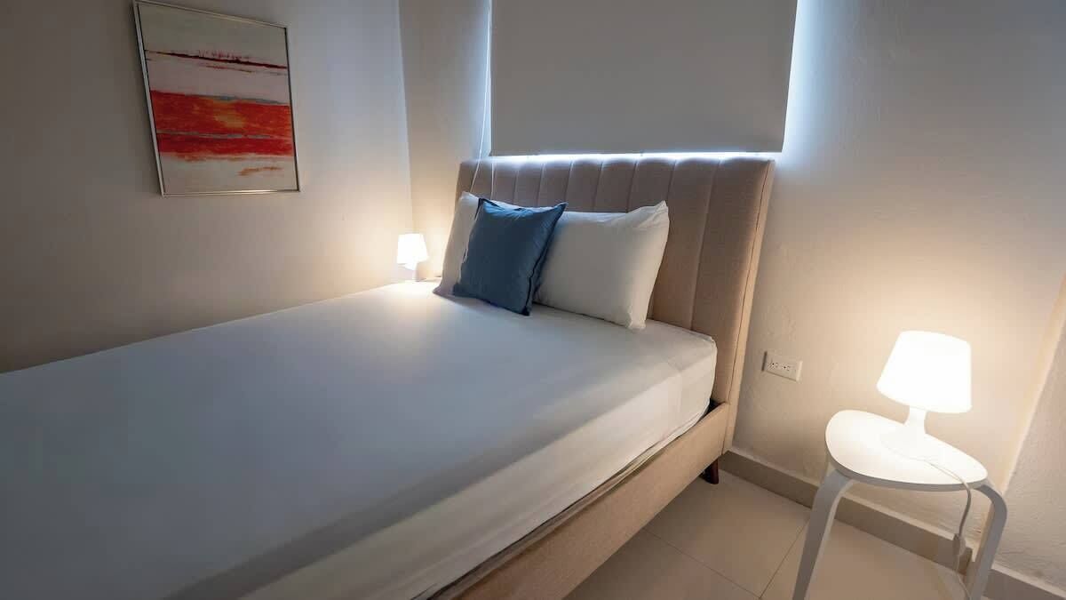 Ein Einzelbett mit Kopfteil, zwei Nachttischlampen, ein kleiner weißer Tisch und ein gerahmtes Bild an der Wand.