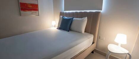 Ein Einzelbett mit Kopfteil, zwei Nachttischlampen, ein kleiner weißer Tisch und ein gerahmtes Bild an der Wand.