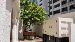 A courtyard with a tree, a building with a white facade, and a large electrical unit. - 3Bedroom Apt. Walking To The Beach C.2 (San Juan)