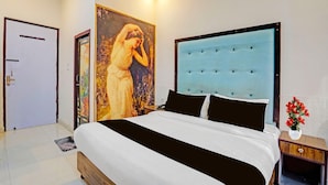 A bedroom with a bed, a nightstand, a door, and a wall painting. - Classic Hotel Haveli inn (Meerut)