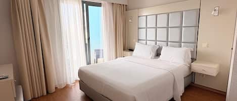 A modern hotel room with a large bed, a bedside table, a nightstand, and a view through a window with curtains.