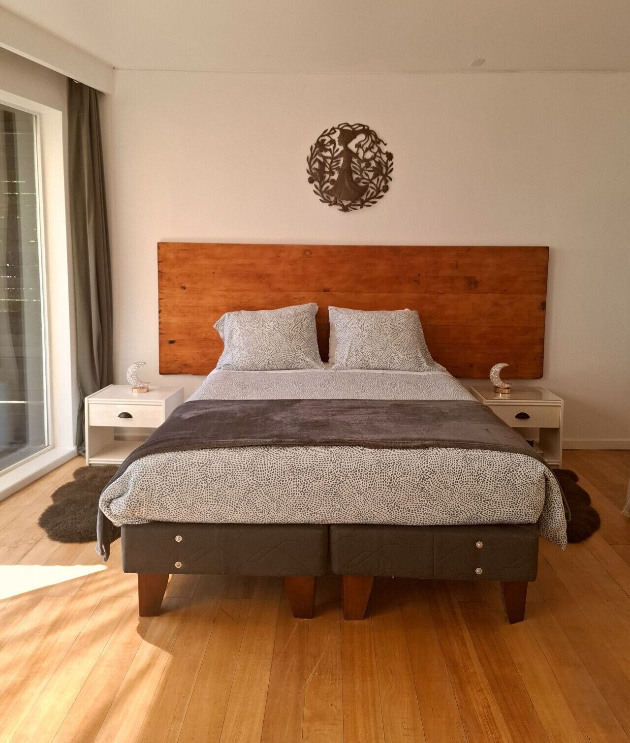Un dormitorio con cabecera de madera, colcha estampada, dos almohadas, mesita de noche con adorno de luna creciente y un ventanal con cortinas.