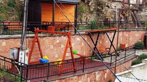 A playground with swings and a small wooden structure, set against a brick wall and a stone building. - Room N 6 by Kraveli (Tbilisi)