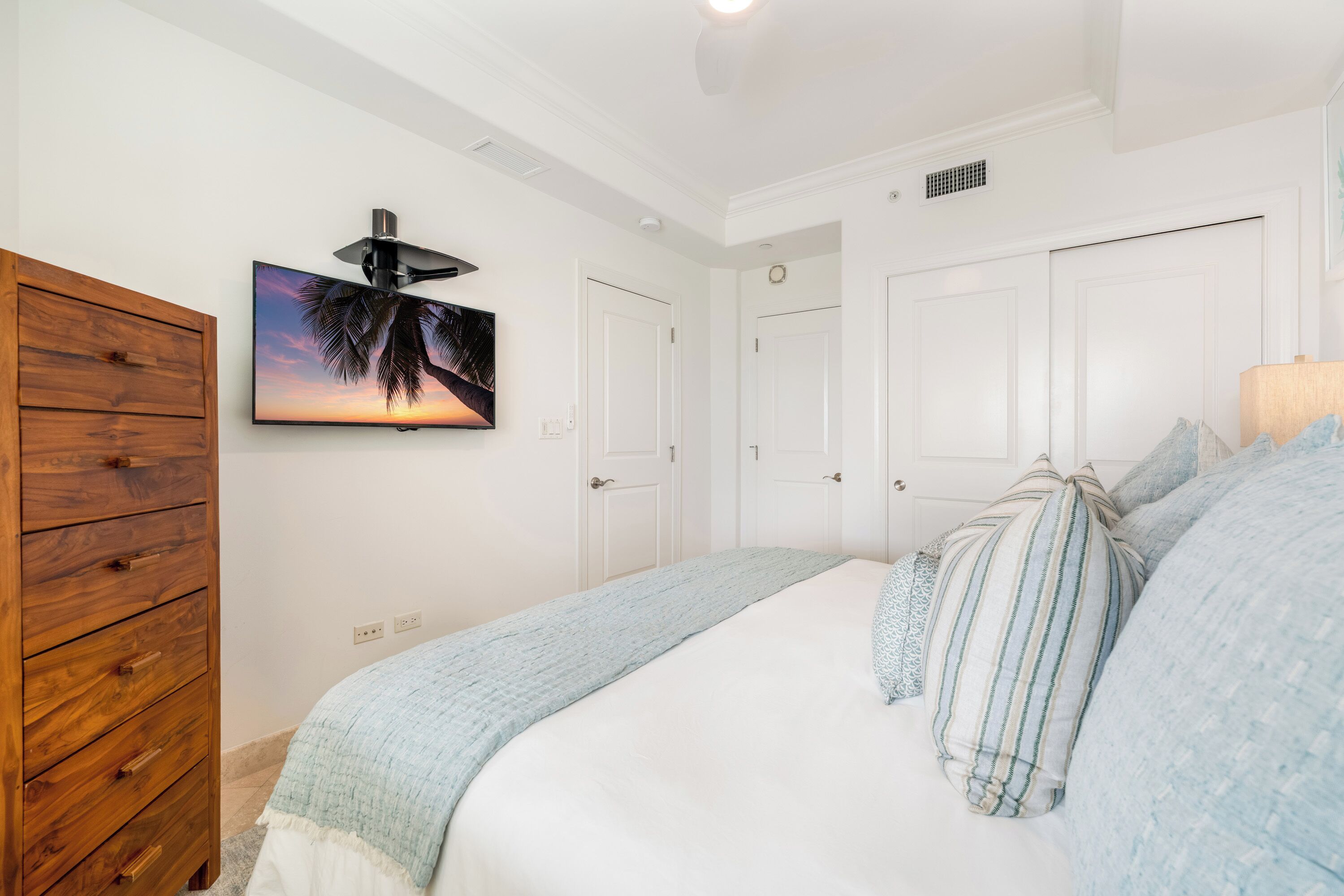 A bedroom with a bed, a wooden dresser, a wall-mounted TV, and white doors.