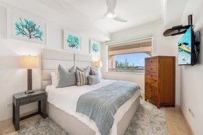 A bedroom with a bed, bedside tables, a wooden dresser, a ceiling fan, and artwork on the walls. - Beachcomber 22 by Grand Cayman Villas (Seven Mile Beach)
