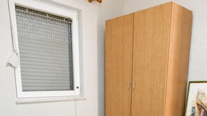 Ein Zimmer mit einem Bett, einem Fenster mit Jalousien, einem Holzschrank und einem gerahmten Bild auf einem kleinen Tisch.