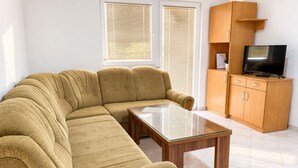 Ein Wohnzimmer mit einer Couch, einem Glastisch, einem Fernseher auf einer Holzkommode und einem Fenster mit Jalousien.